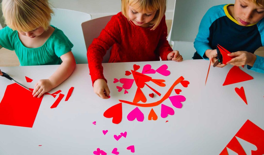 21 adorable Valentine crafts for preschoolers represented by 3 preschoolers cutting paper hearts at a table