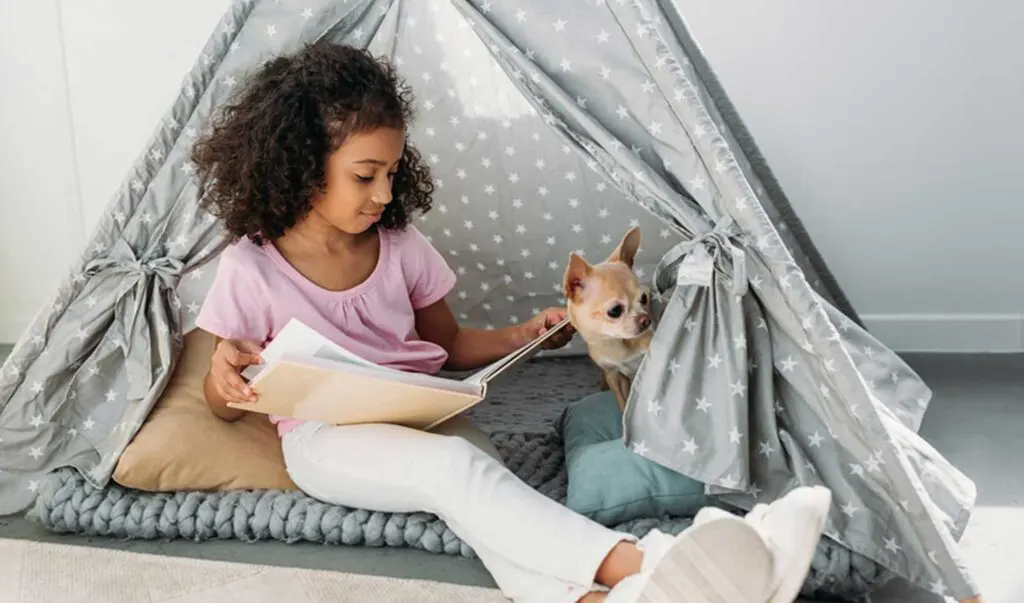 Young African American girl sitting on a floor cushion in a teepee reading nook for kids, reading a book with her Chihuahua sitting next to her.