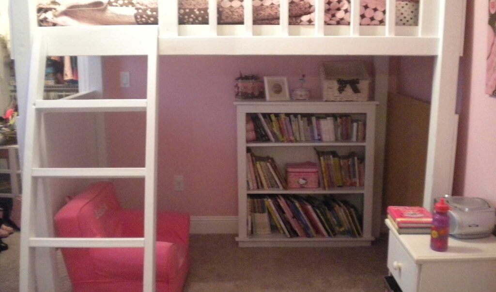 a kids reading nook with hot pink chair and white bookcase set up underneath a white loft bed.