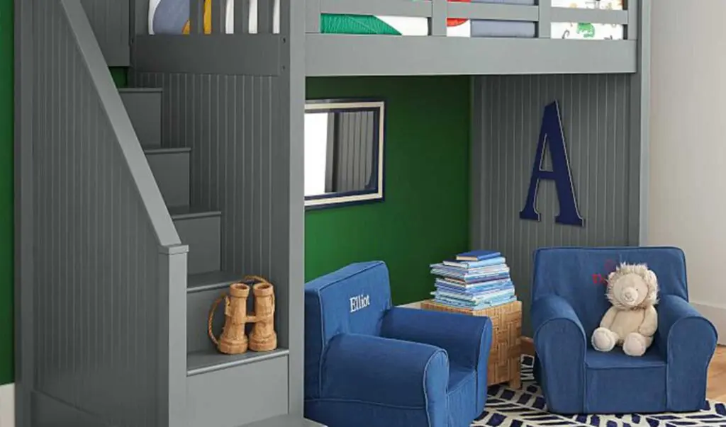 A grey loft bed with enclosed stairs over a book nook with 2 monogrammed blue chairs  and an end table with several books stacked on top of it.