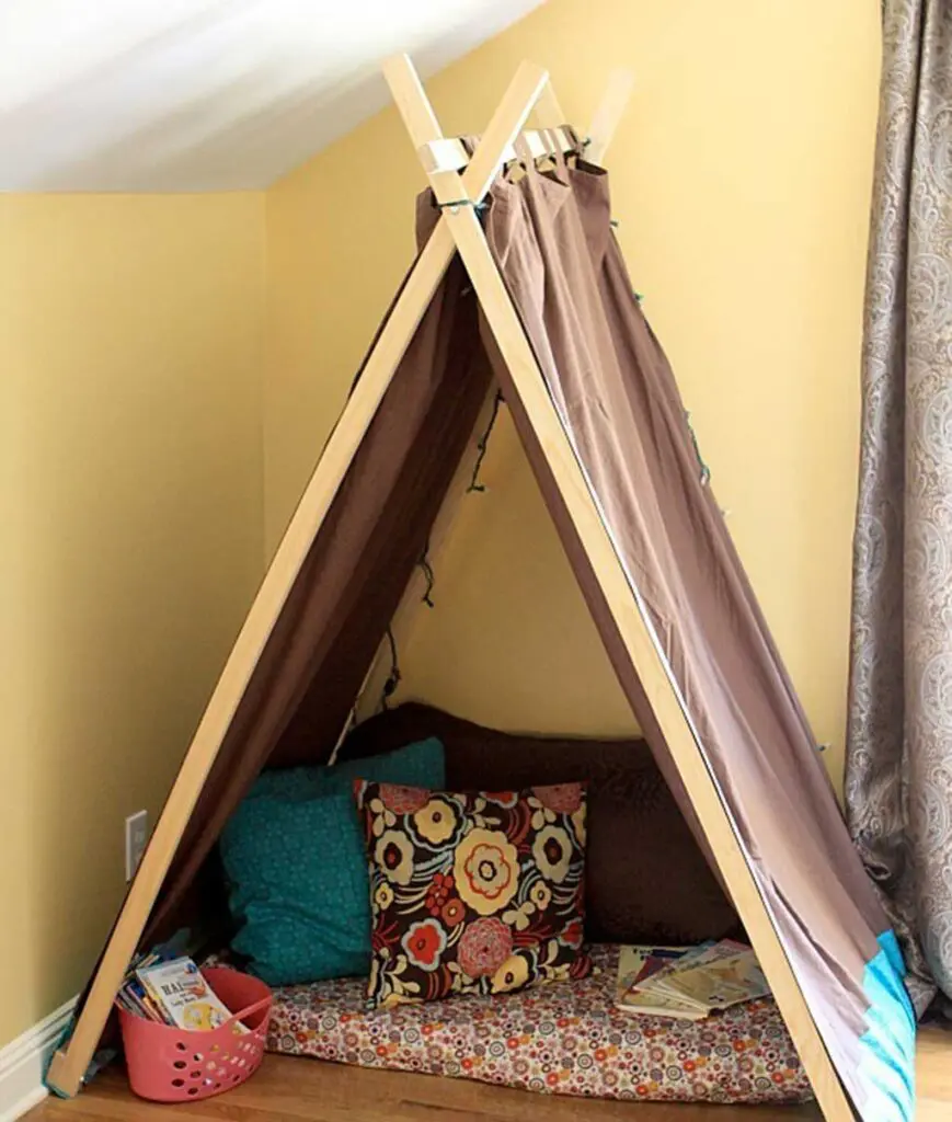 Tee-pee style DIY kids indoor tent with colorful floor mat and throw pillows in abstract floral patterns and a small pink plastic bin holding books.
