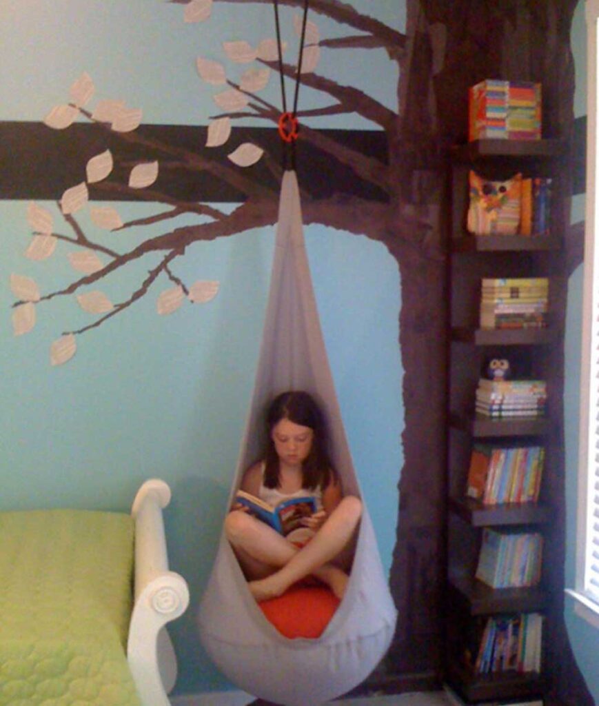 kid sitting in swing chair next to brown bookcase with tree painted on the wall behind it